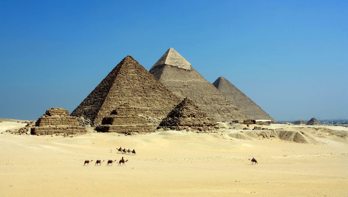 Giza pyramid complex in Egypt. There are two camel trains in front of them, with people on the camels.